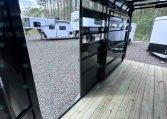 Interior of a glossy black trailer with open side doors, wooden deck, and other horse trailers parked on a gravel lot outside.