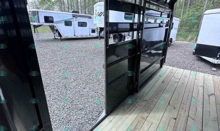 Interior of a glossy black trailer with open side doors, wooden deck, and other horse trailers parked on a gravel lot outside.