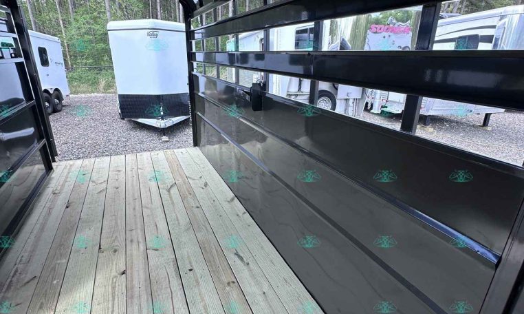 Interior of a black trailer with a wood plank floor and built-in shelves along the right side; other trailers visible outside the open rear door/fan area.