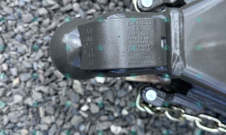 Close-up of a trailer hitch coupler on a gravel surface with safety chains attached nearby.