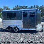 Two-axle gray horse trailer with side double doors and windows, parked on a gravel lot with trees in the background.