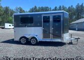 Two-axle gray horse trailer with side double doors and windows, parked on a gravel lot with trees in the background.