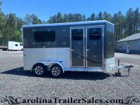 Two-axle gray horse trailer with side double doors and windows, parked on a gravel lot with trees in the background.