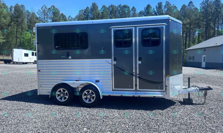 Two-axle gray horse trailer with side double doors and windows, parked on a gravel lot with trees in the background.