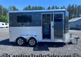 Silver Merhow horse trailer with an open side door, tandem wheels, parked on a gravel lot with trees and a building in the background.