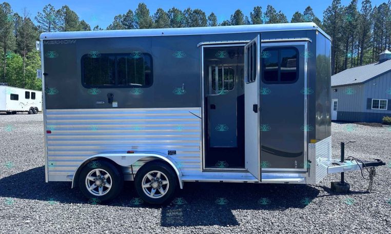 Silver Merhow horse trailer with an open side door, tandem wheels, parked on a gravel lot with trees and a building in the background.