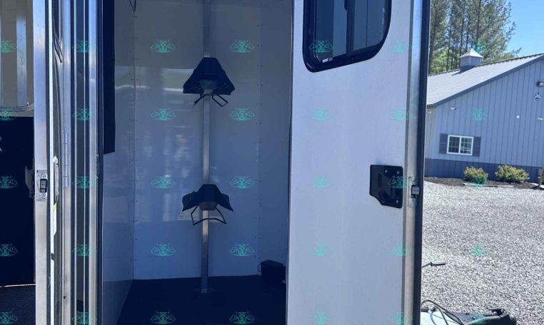 Open rear of an aluminum trailer showing saddle racks along the inner wall; door with window is ajar, parked on a gravel lot with a building in the background.