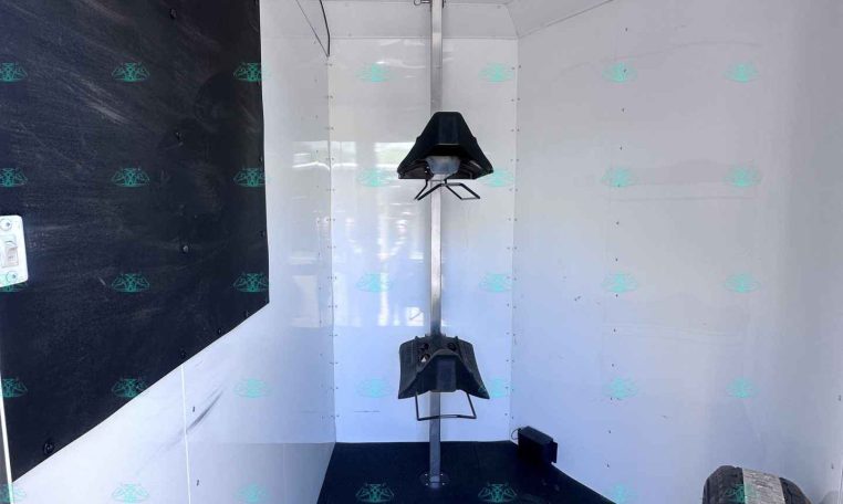 Interior of a light gray trailer with a central vertical support rod and a small black bracket or seat attached, left wall has a black panel with silver D-ring hooks, and a tire is visible in the foreground on the right.