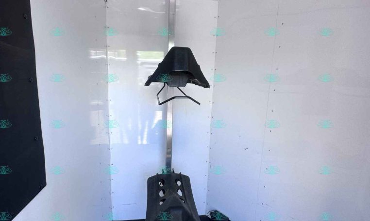 Interior of a white trailer showing a vertical metal rack with a black saddle-like mount attached to it; dark flooring and walls, storage area for tack visible in the space.