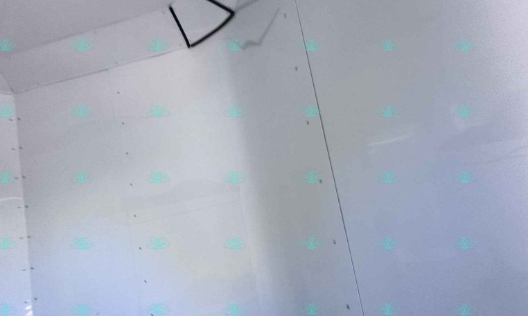 Corner of an unfinished drywall room with taped panel seams and a black metal brace hanging near the ceiling corner; light gray walls and a watermark reading CarolinaTrailersales.com at the bottom.