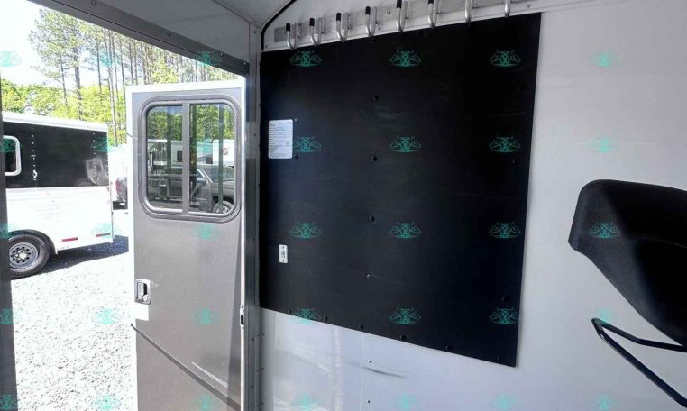 Interior of a white trailer with a row of metal hooks mounted on a wall panel and an open door to the outside outside view of another trailer.