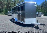 Silver travel trailer on a gravel lot with trees in the background and a second trailer in the distance.