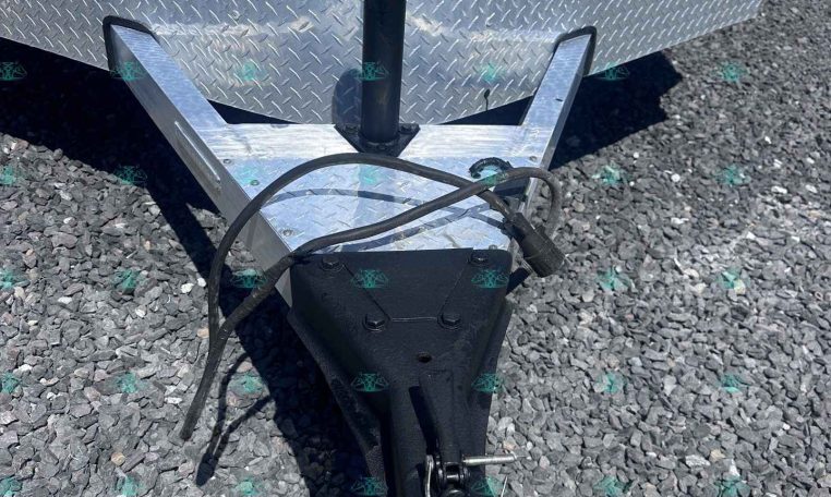 Trailer hitch area: black A-frame tongue, vertical jack, safety chains, and coupler on gravel with a diamond-plate trailer side in the background