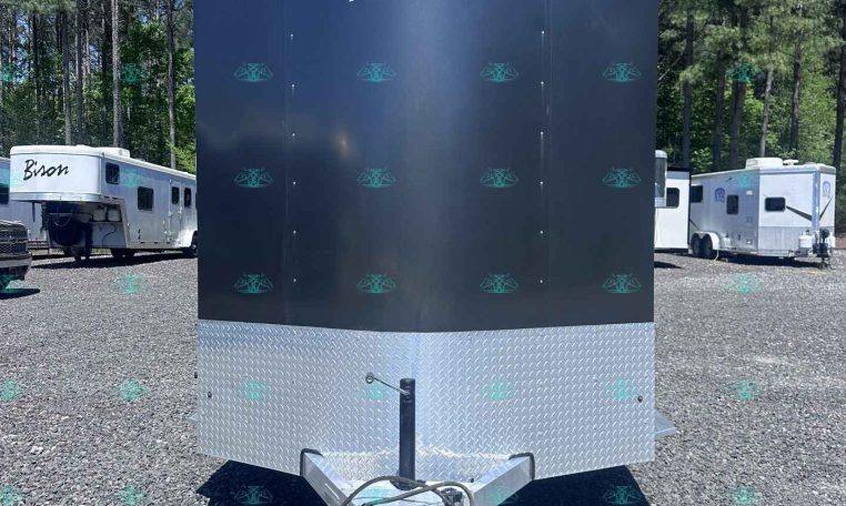 Dark blue Merhow horse trailer with diamond-plate front, attached to a gooseneck hitch, parked in a gravel lot with trees and other trailers in the background.