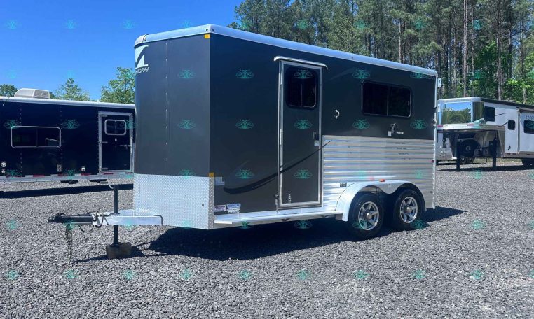 Dark gray tandem-axle trailer on a gravel lot, with trees in the background under a clear blue sky.