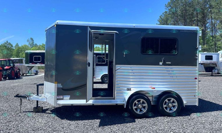 Side view of a dark gray Merhow horse trailer with an open doorway, parked on a gravel lot with trees in the background.