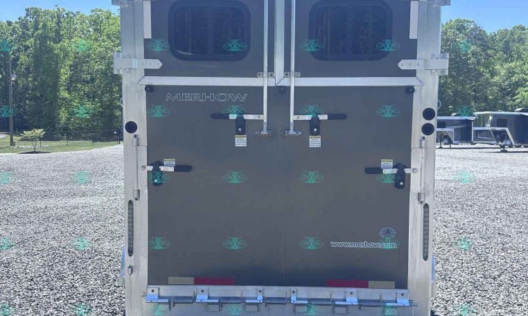 Rear view of a gray Merhow horse trailer with double doors, latches, and small windows on each door, parked on gravel with trees in the background.
