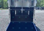 Open horse trailer interior with blue padded stalls and a lowered rear ramp on a gravel lot.