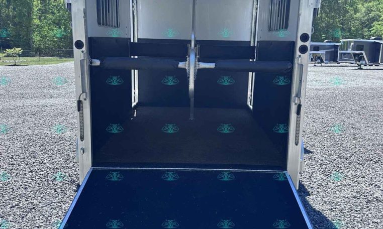 Open horse trailer interior with blue padded stalls and a lowered rear ramp on a gravel lot.
