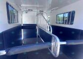 Interior of a white horse trailer with a central divider, blue padded walls, and metal rails dividing stalls, seen from the back ramp area.