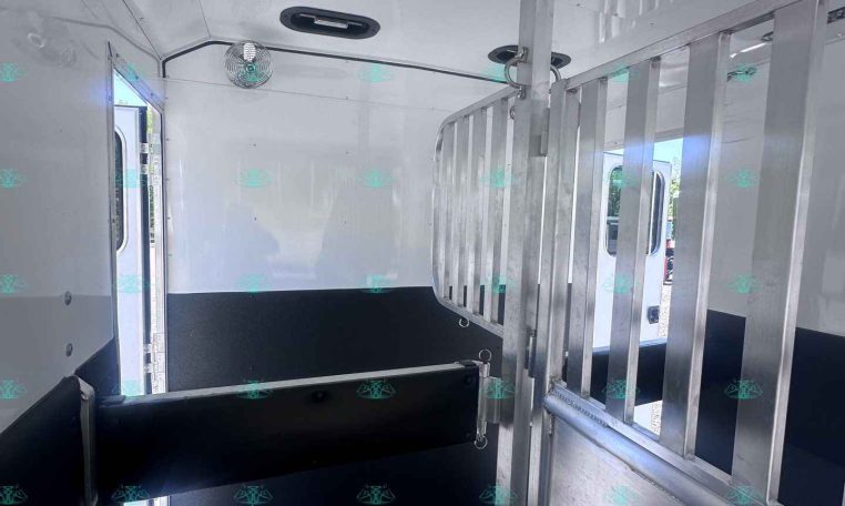 Interior of a horse trailer with a metal vertical divider and a dark non-slip floor, ventilation fan, and light entering from side windows