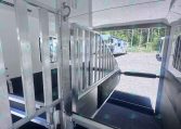 Interior of a metal horse trailer with divider stalls and an open side door showing other trailers outside on a gravel lot.