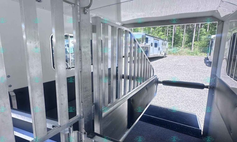 Interior of a metal horse trailer with divider stalls and an open side door showing other trailers outside on a gravel lot.