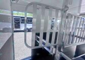 Interior of a metal horse trailer showing a vertical divider gate, loading ramp, and ceiling vent/fan. Outside view of another trailer through the doorway.