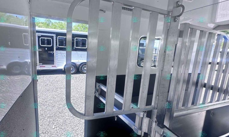 Interior of a metal horse trailer showing a vertical divider gate, loading ramp, and ceiling vent/fan. Outside view of another trailer through the doorway.