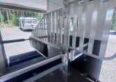 Interior of aluminum horse trailer with stall divider and ramp, viewed from the rear door, outdoors with a parked trailer in the background.