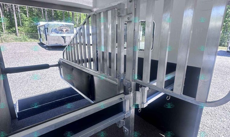 Interior of aluminum horse trailer with stall divider and ramp, viewed from the rear door, outdoors with a parked trailer in the background.