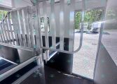 Interior of a metal horse trailer showing a vertical-bar divider gate, hooks and latches, a wall-mounted fan, and a window with outside trailers visible.