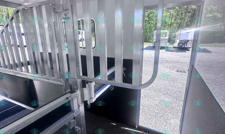 Interior of a metal horse trailer showing a vertical-bar divider gate, hooks and latches, a wall-mounted fan, and a window with outside trailers visible.