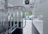Interior of a white livestock trailer with a metal divider gate on the left and a small roof vent; an open door reveals a gravel lot with parked trailers outside.