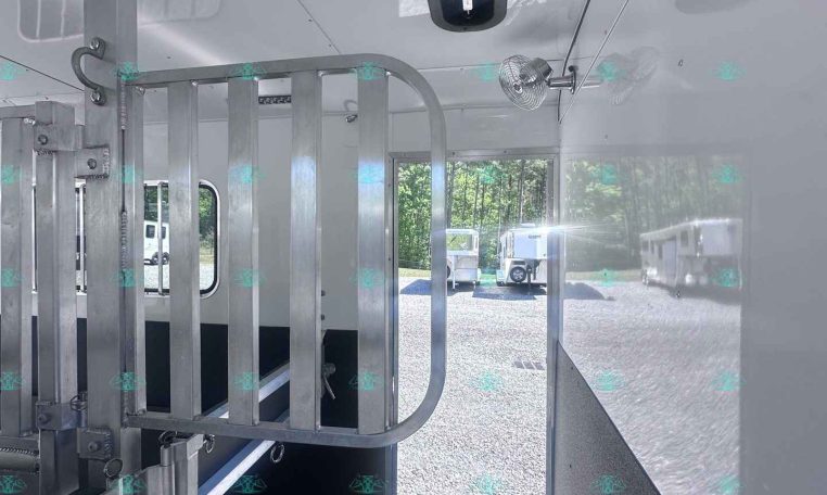 Interior of a white livestock trailer with a metal divider gate on the left and a small roof vent; an open door reveals a gravel lot with parked trailers outside.