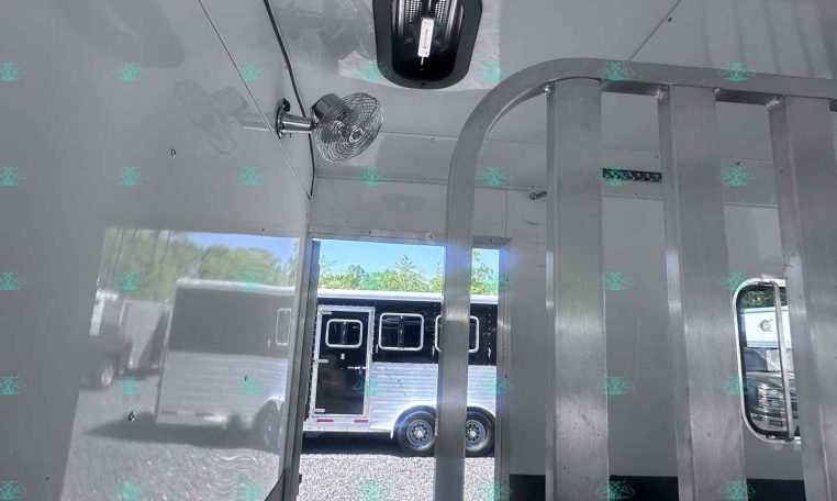 Interior of a light-gray trailer with a metal divider, ceiling vent, and small fan; outside are other trailers.