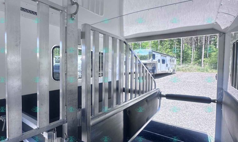 Interior of a metal livestock trailer with aluminum divider gates and a rubberized ramp, seen from the doorway toward an outdoor trailer area