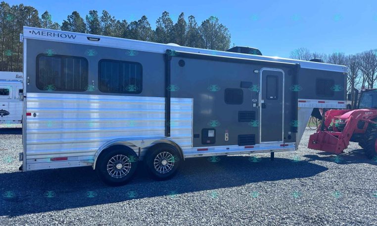 Gray Merhow horse trailer with two axles and side entry door on a gravel lot, with a red tractor nearby in the background.