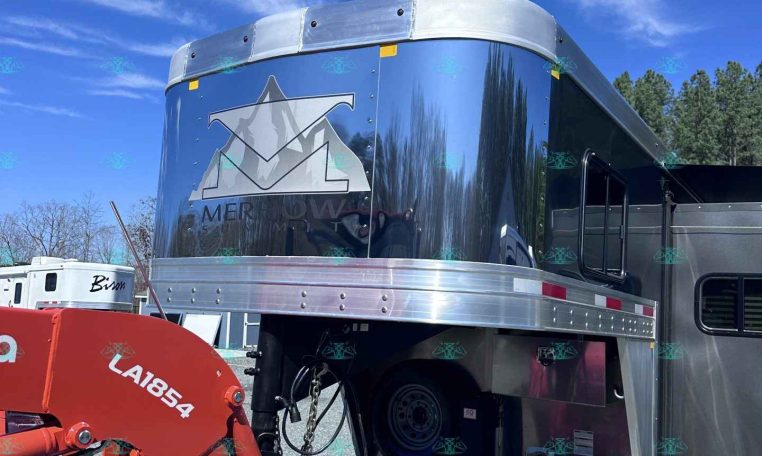 Polished aluminum horse trailer with chrome trim, hitched to a red tractor attachment on a gravel lot.