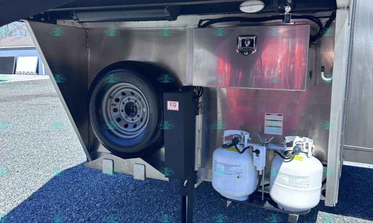 Under-trailer storage area showing a spare tire in the wheel well, a metal utility box, and two connected propane tanks beneath the trailer.