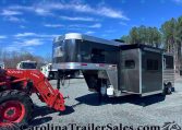 Dark aluminum horse trailer with chrome nose and gooseneck hitch, hitched to a red Kubota tractor in a gravel lot. Trees in the background