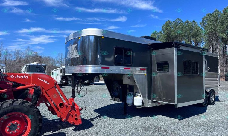 Dark aluminum horse trailer with chrome nose and gooseneck hitch, hitched to a red Kubota tractor in a gravel lot. Trees in the background