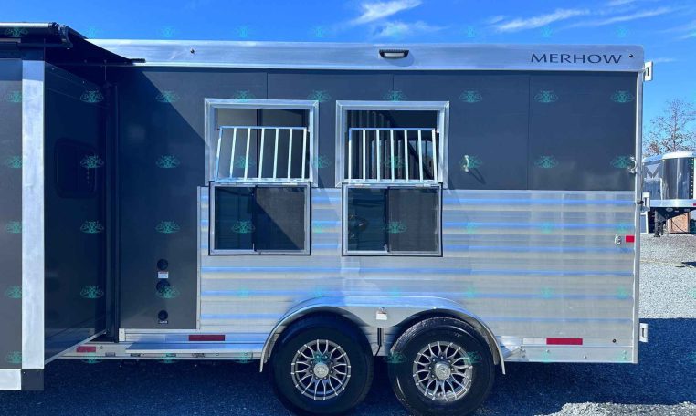 Silver Merhow horse trailer with two barred windows and tandem wheels, parked on a gravel lot under a blue sky.