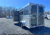 Shiny silver Merhow horse trailer with the side door open, parked on a gravel lot with leafless trees in the background.