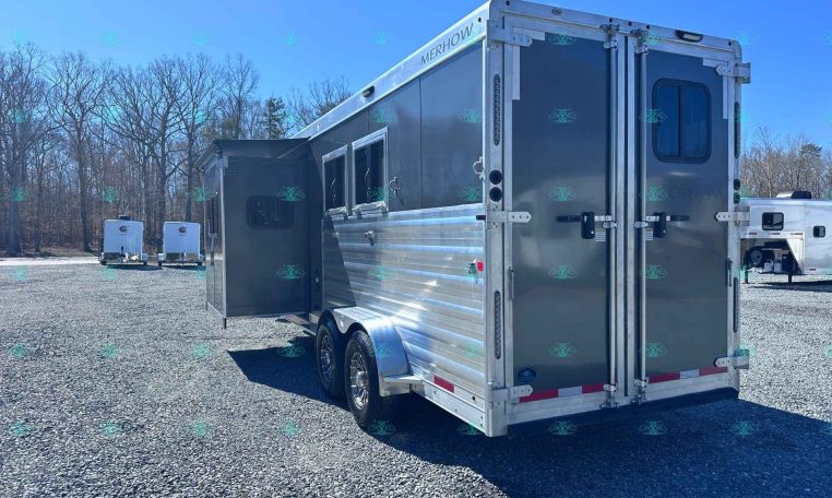 Shiny silver Merhow horse trailer with the side door open, parked on a gravel lot with leafless trees in the background.