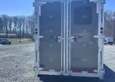 Rear view of a gray enclosed cargo trailer with double doors in a gravel lot; metal latches and a small window on the right door break the monotone surface.