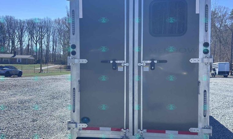 Rear view of a gray enclosed cargo trailer with double doors in a gravel lot; metal latches and a small window on the right door break the monotone surface.