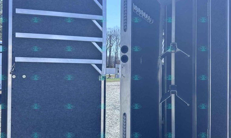 Open trailer doors reveal an interior with hanging racks, hooks, and storage poles inside a gravel lot.