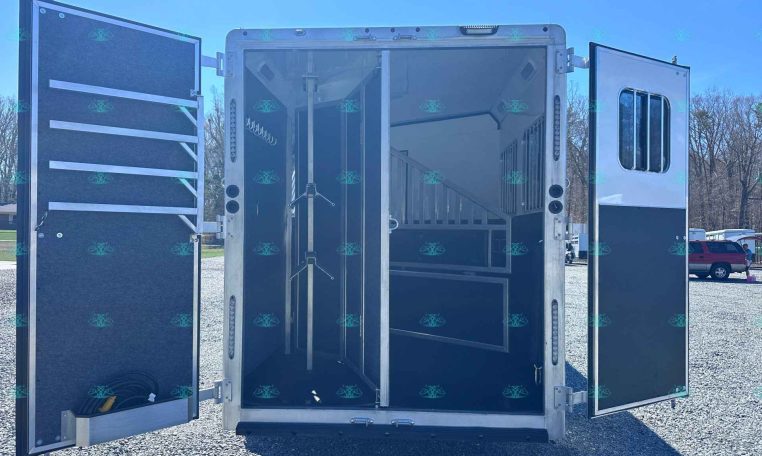 Open horse trailer with interior stalls and divider, doors swung wide, parked on gravel.