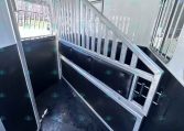 Interior of a horse trailer showing a metal divider with vertical bars and a padded wall along the stall, forming a narrow aisle with a wet, rubberized floor.